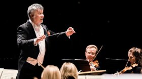 Conductor of a symphony orchestra holding a baton with outstretched arms over the musicians seated in front of him.