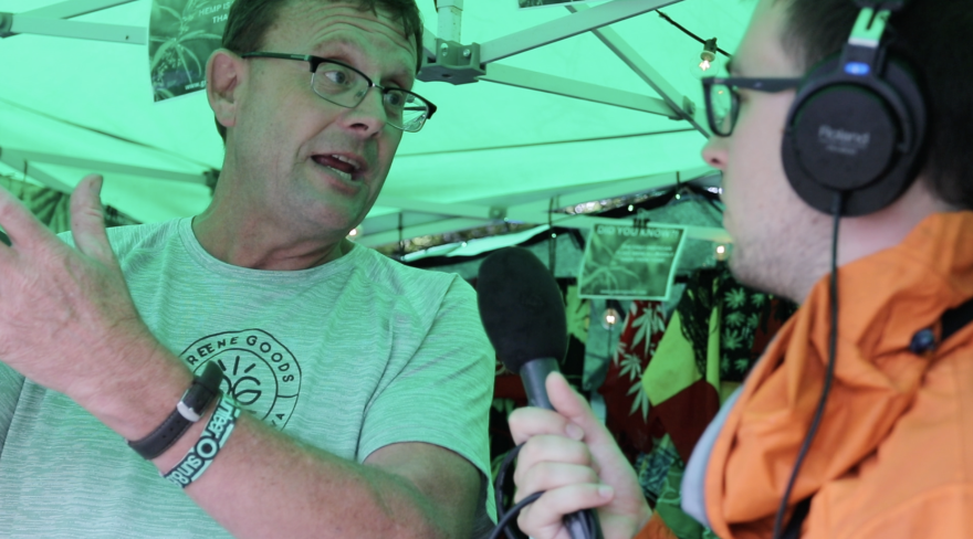  Andy Krieger with Greene Goods Market and Greenhouses at Hinterland in August 2023 talks with IPR's Phineas Pope in his vendor booth. He's wearing a green shirt with his company's logo on the front. 