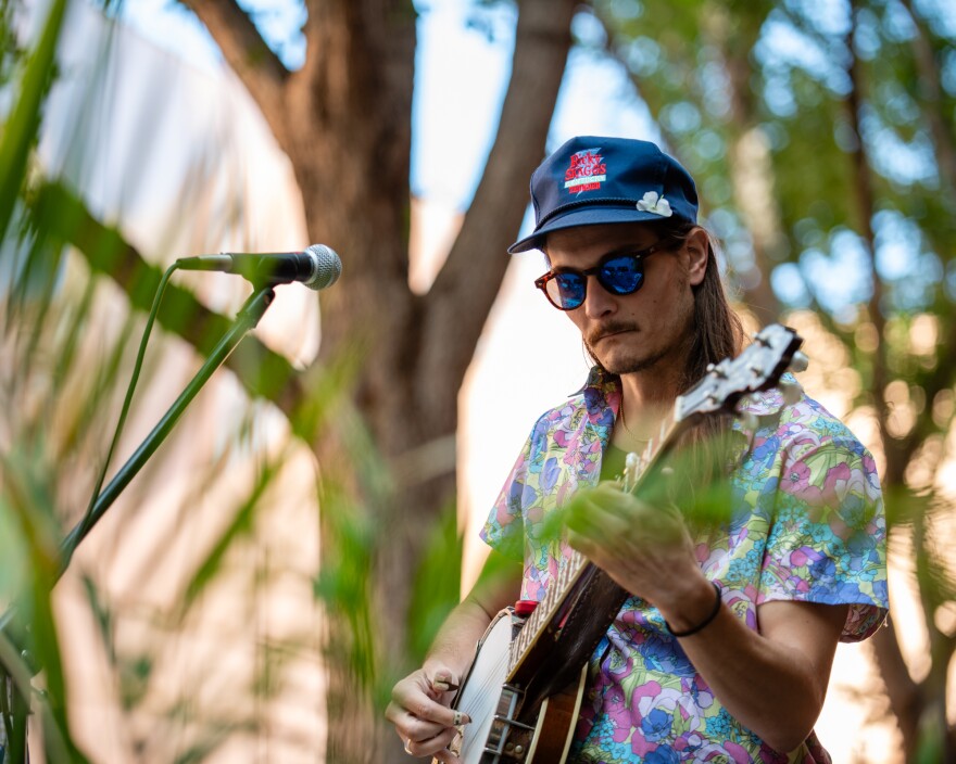 with greenery in the foreground, a man plays a banjo in a Hawaiian shirt