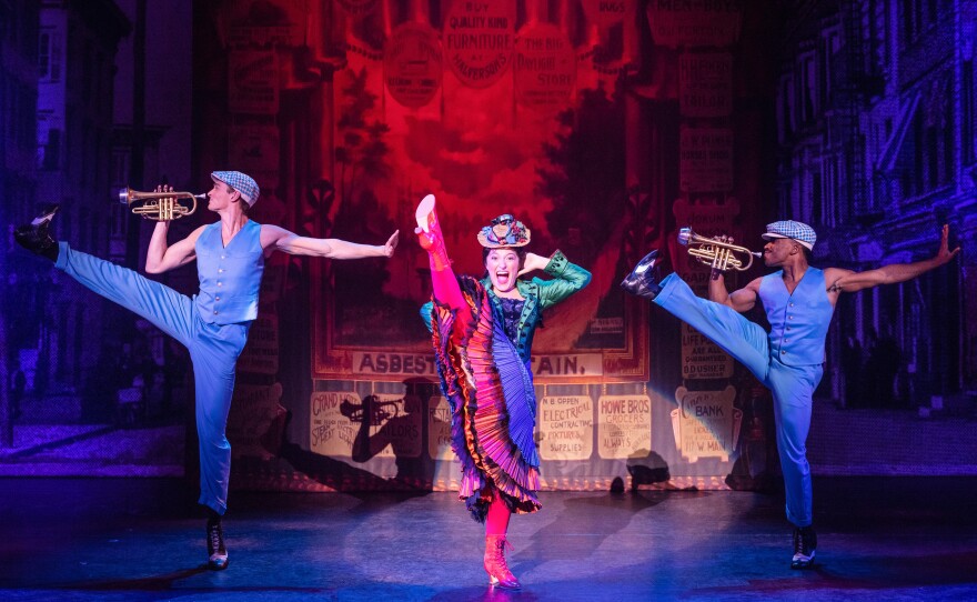 An actress sings on stage next to two trumpet players.