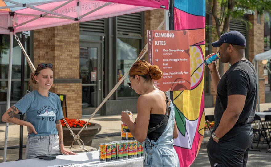 A man and a woman stand facing another women who is inside a vendor booth at the 2023 Des Moines Arts Festival. The couple is drinking Climbing Kites social beverage. 