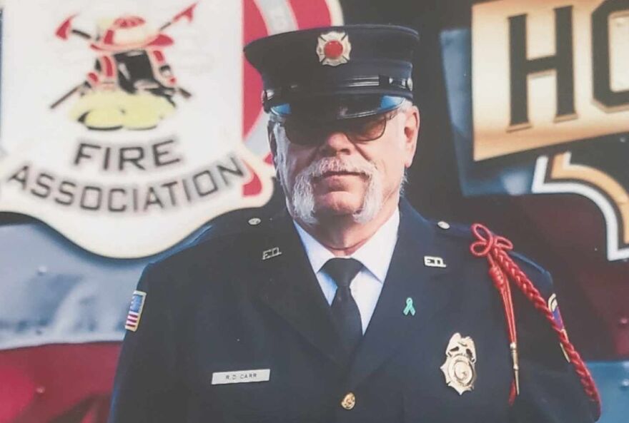 Photo of a firefighter in front of a fire association logo. He is wearing official gear and has dark glasses and facial hair.