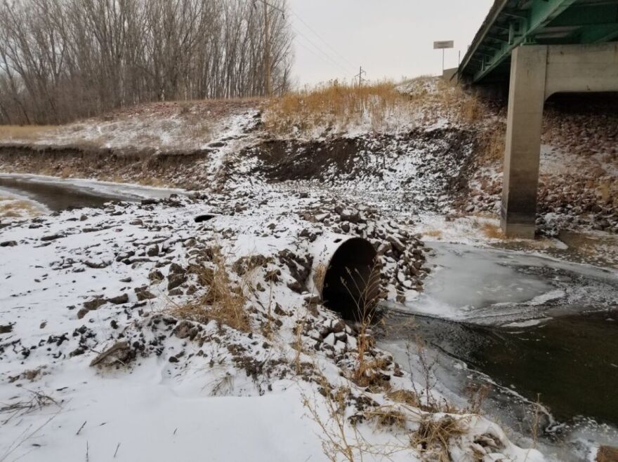 A pipe sticks of the frozen grounds with a bit of snow. Water flows from the pipe underneath a road bridge.