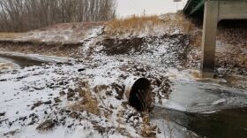 A pipe sticks of the frozen grounds with a bit of snow. Water flows from the pipe underneath a road bridge.