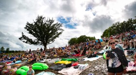 Thousands sit on towels on a muddy hill. There is one big tree and more clouds than sky.