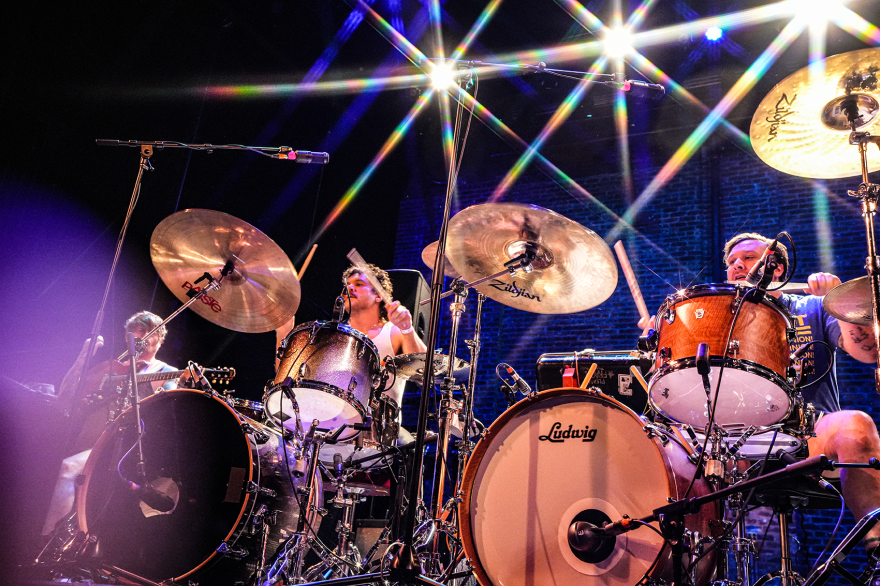 Two drums kits are set on stage next to each other with two men playing them in unison