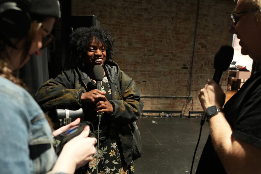  A woman holding a microphone wearing a floral dress and a jacket speaks with a man and a woman also holding microphones