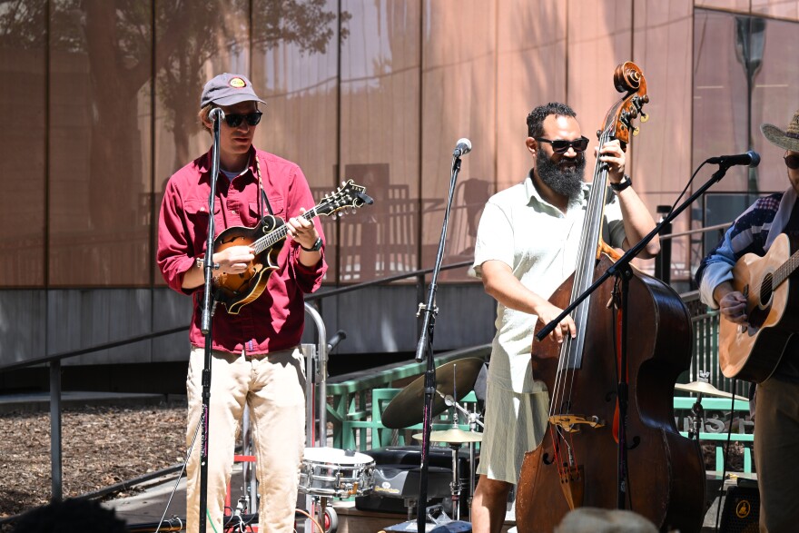 A man in a red shirt and khaki pants plays mandolin next to a Black man playing a stand up bass. 