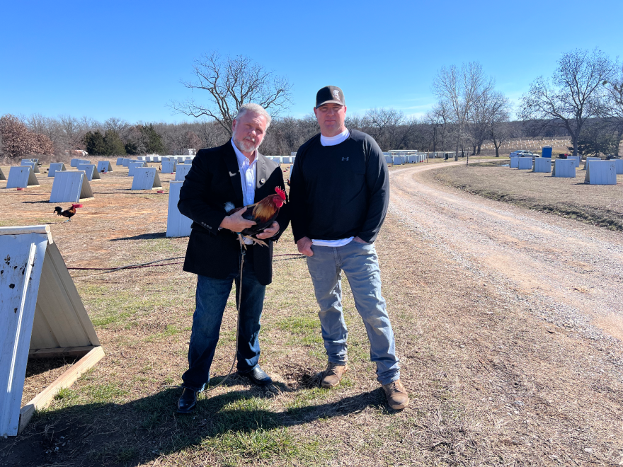 A man in a blazer stands with a rooster next to a man in a ball cap in front of rows of white A-frame shelters for roosters.