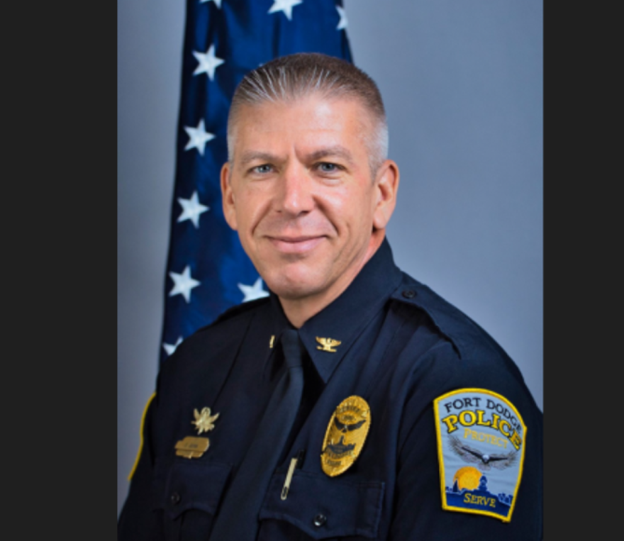 A man is smiling and wearing a police officer's uniform with a badge and a patch on his arm that says Fort Dodge Police.