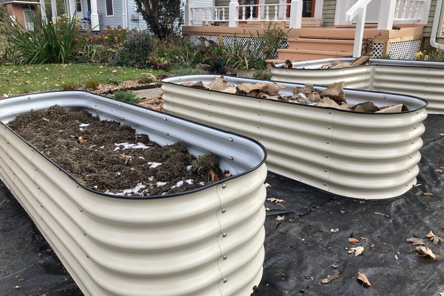 Three off-white metal garden beds filled with soil, frost, and cardboard rest atop a black tarp outside a house. 