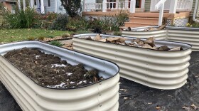 Three off-white metal garden beds filled with soil, frost, and cardboard rest atop a black tarp outside a house. 