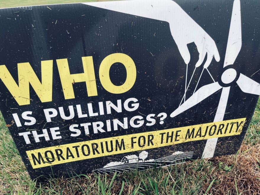 Sign with the words "Who is pulling the strings? Moratorium for the majority." The background is back with yellow and white text with a windmill with puppet strings and a hand. On the bottom is a farm field scene.