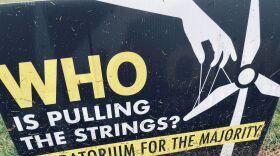 Sign with the words "Who is pulling the strings? Moratorium for the majority." The background is back with yellow and white text with a windmill with puppet strings and a hand. On the bottom is a farm field scene.