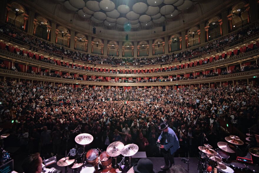 A band plays on stage to a full auditorium, many in the crowd are standing and facing the stage.