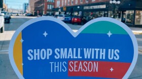 A multi-colored heart of yellow, blue, red, and blac, is in the foreground of a city street. The heart says "shop small with us this season."