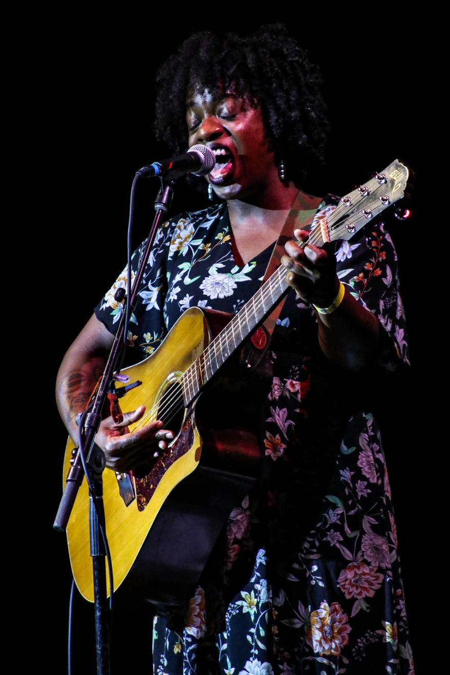 A woman in a black floral dress plays the guitar and sings.