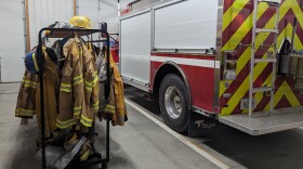 Picture of inside a fire station with uniforms to the left and a fire truck to the right.