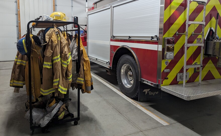 Picture of inside a fire station with uniforms to the left and a fire truck to the right.