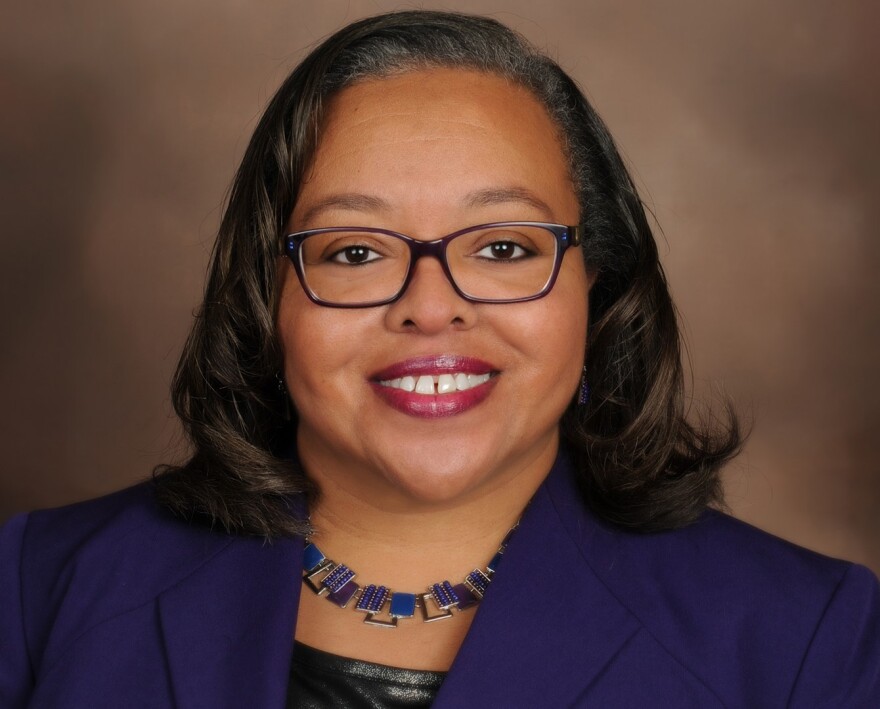 A lady with dark hair and eyes is wearing glasses and wearing a blue blazer and necklace. 