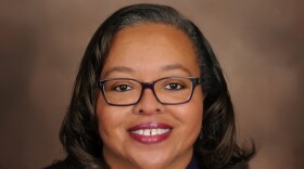 A lady with dark hair and eyes is wearing glasses and wearing a blue blazer and necklace. 