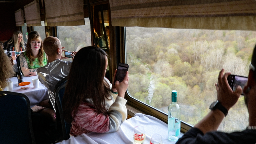 People sit at dining tables with white table cloths in a train dining car. They look out windows at a valley in spring in Iowa. 