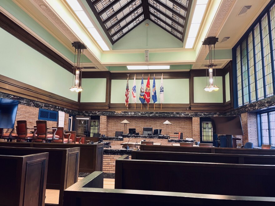 Photo of a courtroom with dark brown wooden benches with brick walls and swirled marble inlays. There are big stained-glass windows to the right. The whole room looks like it was designed by Frank Lloyd Wright.