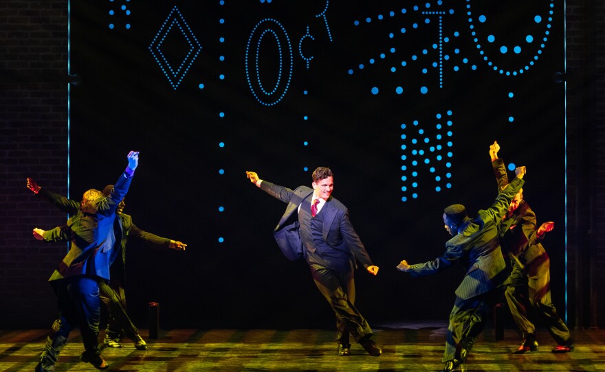 Five men in suits dance on stage in front of neon lights.