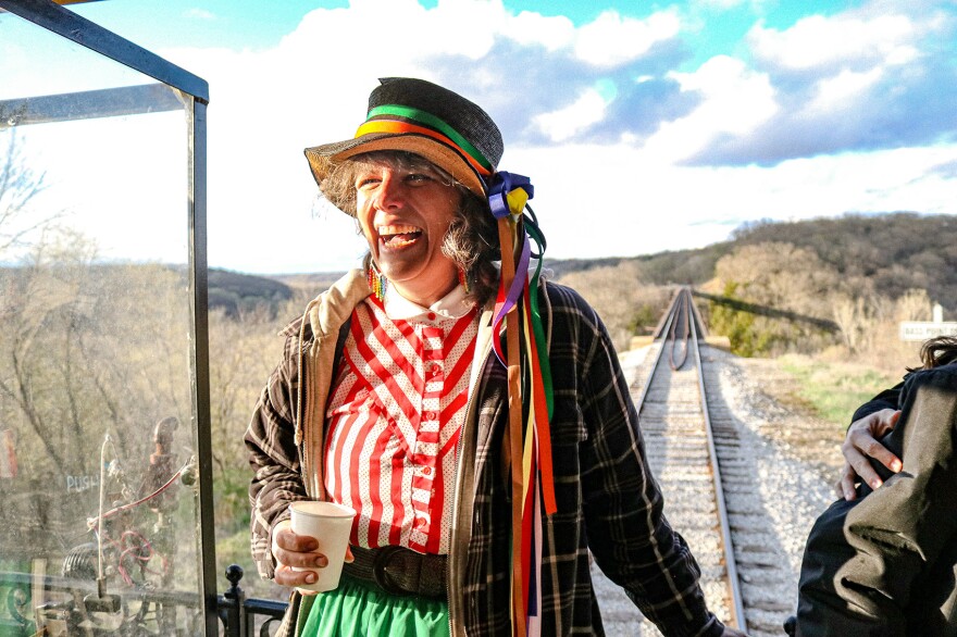 A woman wearing a hat with ribbons tied around it stands on the caboose of a train. Above her, blue sky, behind her, a railroad track in spring. 