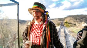 A woman wearing a hat with ribbons tied around it stands on the caboose of a train. Above her, blue sky, behind her, a railroad track in spring. 