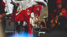 Orville Peck, dressed in red pants and a red fringe shirt and a cowboy hat, plays his guitar on the Hinterland mainstage. 