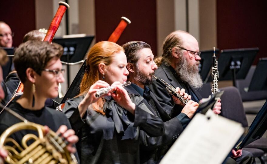 Four people are playing instruments. There is a lady holding a French horn, another lady with a flute, and two men with oboes. 