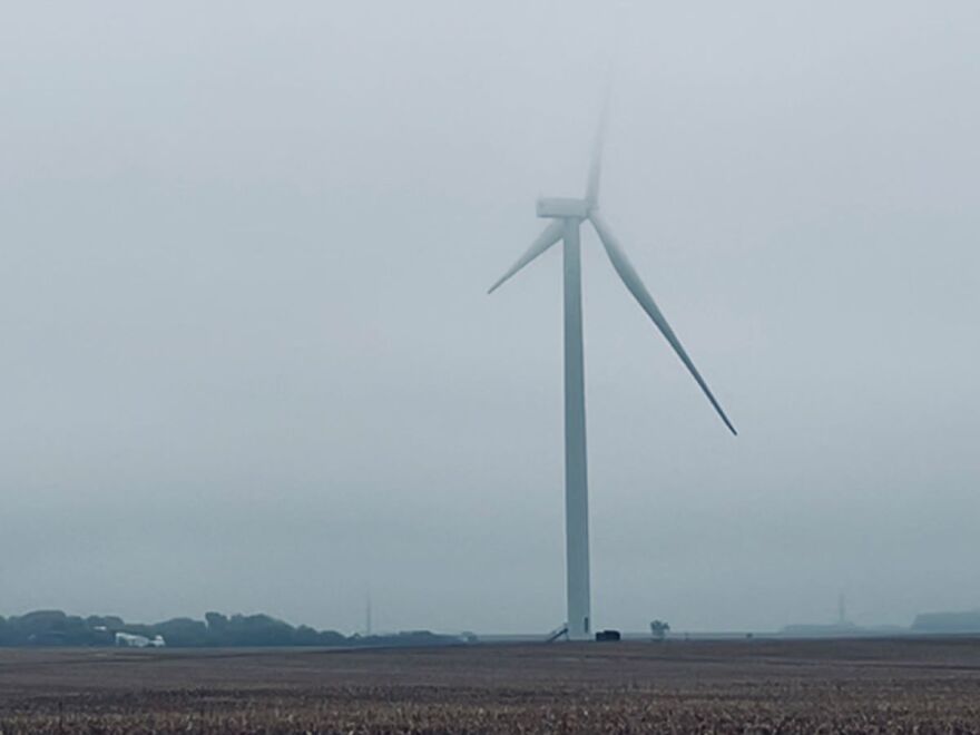 Image shows a white white turbine sitting in the middle of a harvested corn field. The air is hazy due to fog.