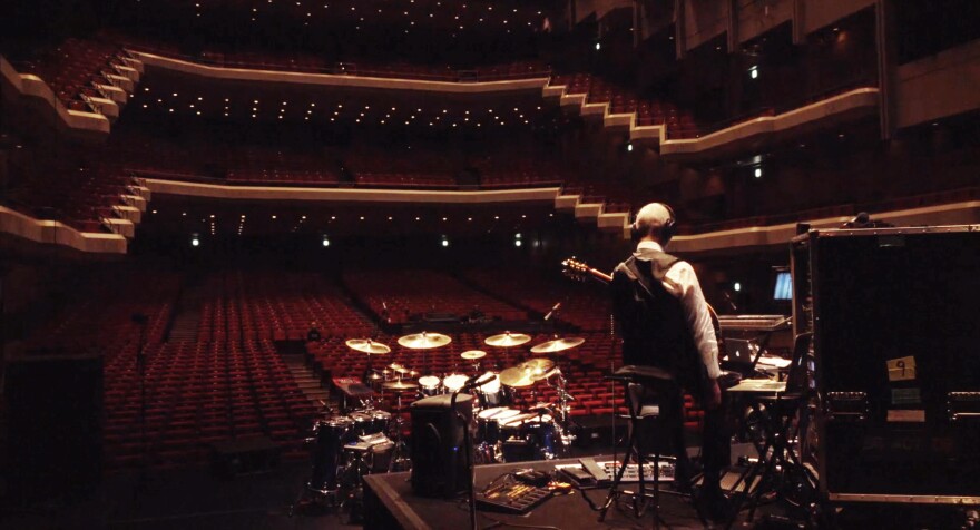 A man sits alone on a stool on stage with a guitar, staring out into an empty auditorium.